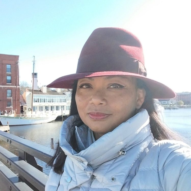Portrait of Genevieve Cheung wearing a hat and winter coat by the water during travel, reflecting a moment of observation and presence.