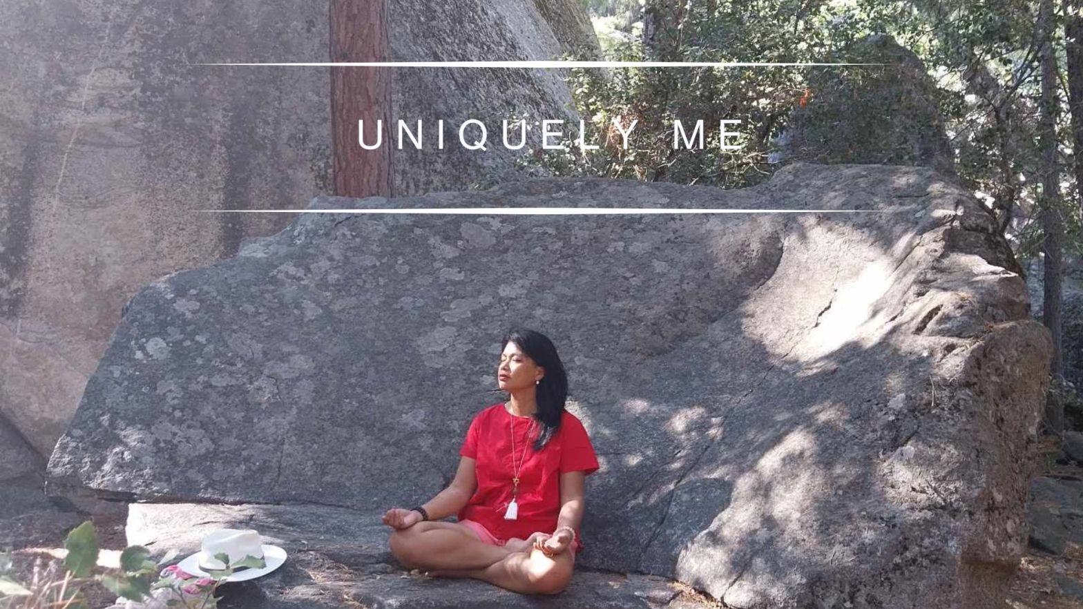 Genevieve Cheung sitting calmly on a large stone in nature, wearing a red dress, eyes closed in quiet presence, surrounded by trees and natural light.