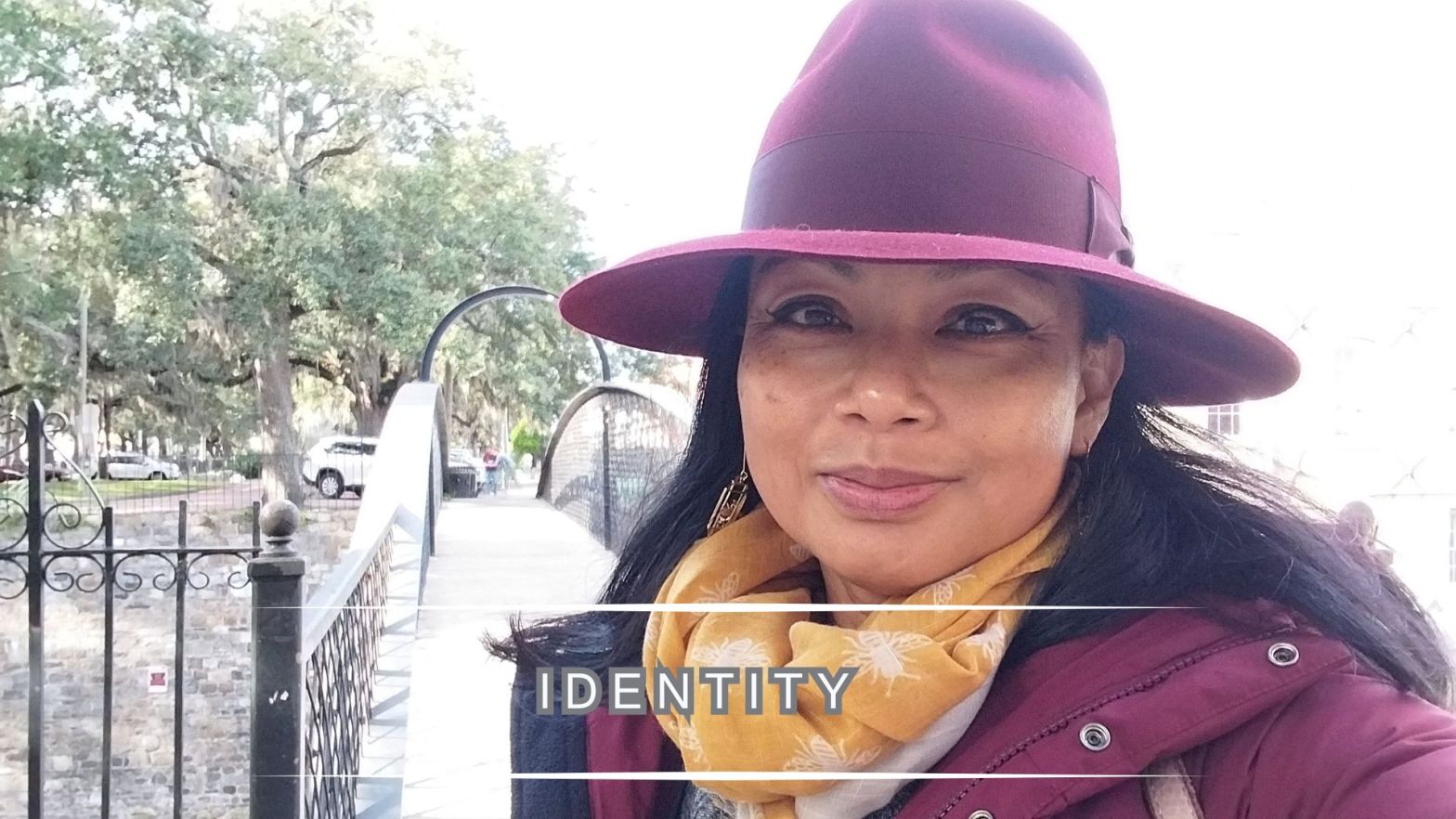 Genevieve Cheung wearing a burgundy hat and yellow scarf, standing on a small bridge outdoors, with the title “Identity” overlaid on the image.