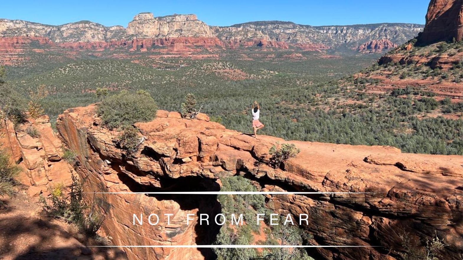 Genevieve Cheung standing in tree pose on a red rock cliff overlooking a vast desert canyon landscape under a wide blue sky, symbolizing inner steadiness in uncertain times.